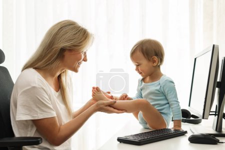 Téléchargez les photos : Jeune mère indépendante souriante joue avec son petit fils assis sur le bureau à son lieu de travail à la maison. - en image libre de droit