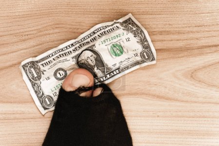 Photo for Closeup shot of a male foot in a black hoaly sock with sticking out toe and a one dollar banknote on wooden floor background. Concept of poverty and financial crisis. - Royalty Free Image
