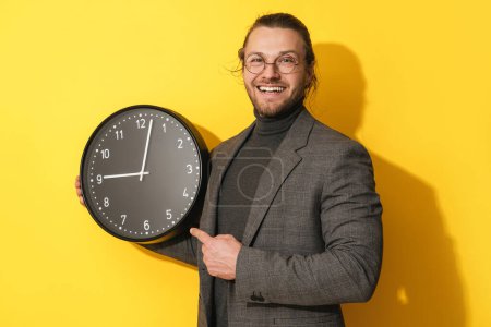 Téléchargez les photos : Homme barbu joyeux portant des lunettes tenant une grande horloge sur fond jaune - en image libre de droit