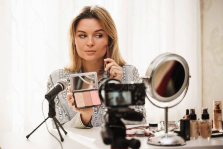 Photo for Woman beauty blogger applying blush on her face during video tutorial recording for her followers - Royalty Free Image
