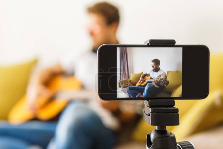 Foto de Un joven músico blogger con gafas sentado en un sofá frente a una cámara está tocando casualmente la guitarra acústica en una habitación iluminada. - Imagen libre de derechos