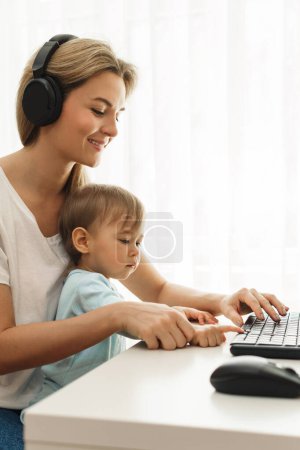 Téléchargez les photos : Jeune mère pigiste travaille sur ordinateur à son bureau à la maison avec son petit fils assis sur ses genoux. - en image libre de droit