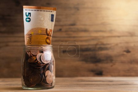 Foto de Primer plano de un billete de 50 euros dentro de un frasco de vidrio lleno de monedas de cobre con un fondo de pared de madera. - Imagen libre de derechos