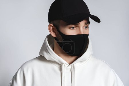 Foto de Joven con capucha blanca, gorra de béisbol negra y mascarilla de tela contra fondo gris - Imagen libre de derechos