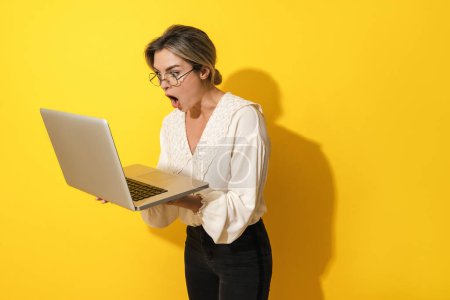 Photo for Young suprised woman wearing eyeglasses using laptop computer on yellow background - Royalty Free Image
