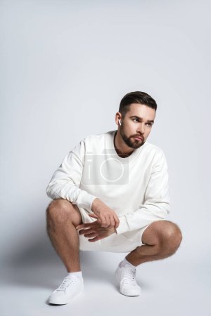Téléchargez les photos : Beau homme portant un sweat-shirt blanc et un short avec des écouteurs sans fil assis sur fond gris - en image libre de droit
