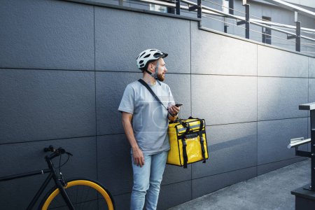 Téléchargez les photos : Jeune messager express de livraison de nourriture avec sac isolé sur le côté tient le téléphone près du vélo garé. - en image libre de droit