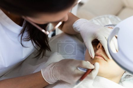 Téléchargez les photos : Maquilleuse professionnelle permanente et sa cliente pendant la procédure de rougissement des lèvres - en image libre de droit