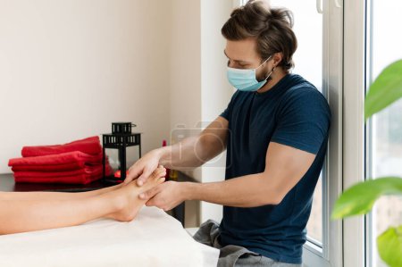 Téléchargez les photos : Masseur professionnel homme portant un masque de prévention pendant la séance de massage des pieds féminins - en image libre de droit