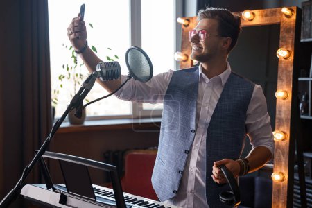 Photo for Young smiling singer and pianist is taking a selfie with his phone at a home music studio in front of a piano with a makeup mirror behind his back. - Royalty Free Image