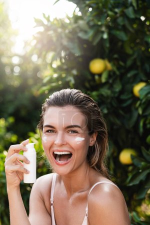 Photo for Outdoors portrait of joyful and beautiful woman applying moisturizing cream or sunblock on her facial skin - Royalty Free Image