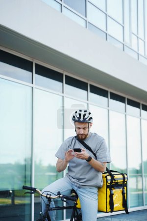 Photo for Young smiling express food delivery courier riding bicycle with insulated bag behind his back is looking at his phone. - Royalty Free Image
