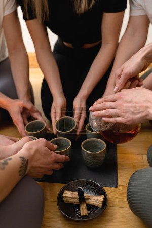 Photo for Young woman is pouring hot tea to her friends sitting around a tray during a tea ceremony. - Royalty Free Image