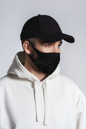Photo for Young man wearing white hoodie, black baseball cap and cloth face mask against gray background - Royalty Free Image