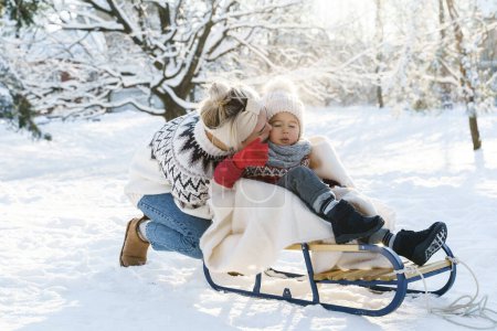 Foto de Joven madre y su lindo hijo pequeño con trineo retro en un parque nevado durante el soleado día de invierno - Imagen libre de derechos