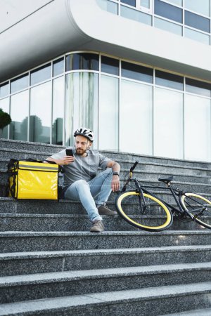 Photo for Young express food delivery courier is sitting on the stairs with insulated bag and bicycle and looking at his phone. - Royalty Free Image
