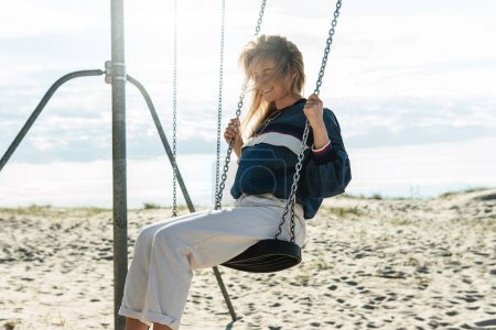 Photo for Young pretty woman is swinging on the swing and smiling at the sandy beach on a sunny day. - Royalty Free Image