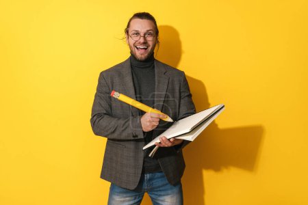 Photo for Cheerful bearded man wearing glasses holding big pencil and notebook on yellow background - Royalty Free Image
