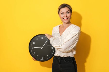 Photo for Young funny woman with silly facial expression holding big clock on yellow background - Royalty Free Image