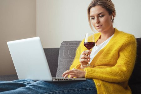 Téléchargez les photos : Jeune femme assise sur le canapé buvant des gémissements rouges en regardant un film ou en travaillant sur son ordinateur portable à la maison - en image libre de droit