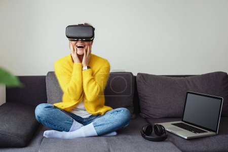 Téléchargez les photos : Jeune femme excitée assise sur le canapé et utilisant un casque de réalité virtuelle à la maison - en image libre de droit