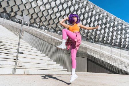 Téléchargez les photos : Femme active insouciante danseuse portant des vêtements de sport colorés s'amusant dans la rue pendant la journée ensoleillée - en image libre de droit