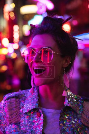 Photo for Young stylish woman wearing jacket with shining sequins on the city street with neon lights - Royalty Free Image