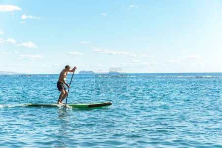 Photo for Young male surfer is riding a standup paddleboard and rowing with a paddle in ocean - Royalty Free Image