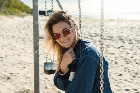 Téléchargez les photos : Jeune jolie femme est assise sur la balançoire et souriant à la plage de sable. - en image libre de droit