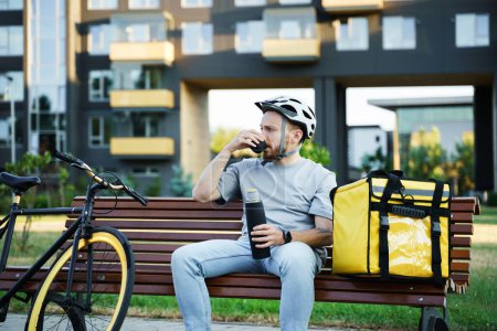 Téléchargez les photos : Jeune messager de livraison express boit du café tout en étant assis sur un banc avec sac isolé et vélo à proximité. - en image libre de droit