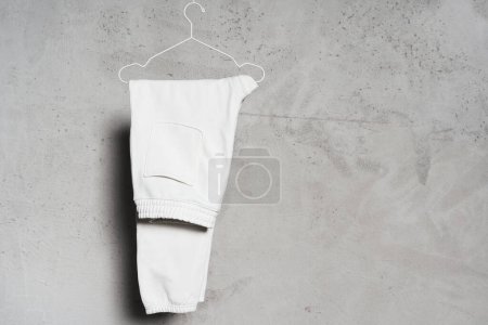 Photo for Mockup of blank white sweatpants hanging on the thin metallic hanger against light concrete wall - Royalty Free Image