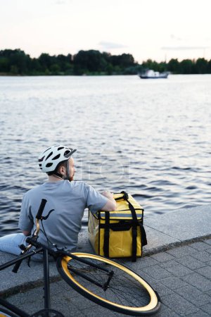 Foto de Joven mensajero entrega express está sentado en un muelle y mirando el agua del río tranquilo con bolsa aislante y bicicleta cerca. - Imagen libre de derechos