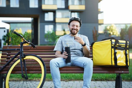 Téléchargez les photos : Jeune courrier de livraison express riant verse du café tout en étant assis sur un banc avec sac isolé et vélo à proximité. - en image libre de droit