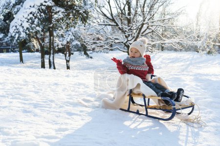 Photo for Cute toddler boy sitting on the sleigh in a snowy city park during sunny winter day - Royalty Free Image