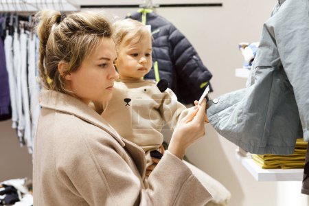 Téléchargez les photos : Jeune femme avec son bébé fils choisir des vêtements dans le magasin de vêtements - en image libre de droit