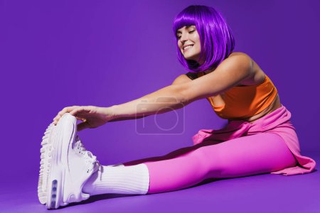Téléchargez les photos : Jeune femme joyeuse portant des vêtements de sport colorés pendant l'entraînement d'étirement sur fond violet - en image libre de droit