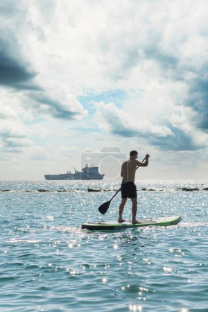 Photo for Young male surfer is riding a standup paddleboard and rowing with a paddle in an ocean with a ship on background. - Royalty Free Image