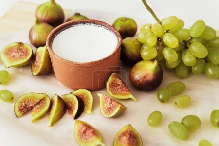 Foto de Delicioso yogur griego natural en tazón de barro con higos y uva sobre papel pergamino blanco - Imagen libre de derechos