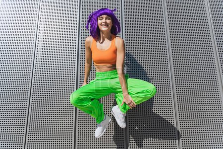 Foto de Mujer joven despreocupada con ropa deportiva de colores saltando en la calle durante el día de verano - Imagen libre de derechos