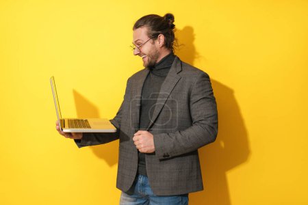 Téléchargez les photos : Beau homme barbu heureux portant des lunettes à l'aide d'un ordinateur portable sur fond jaune - en image libre de droit