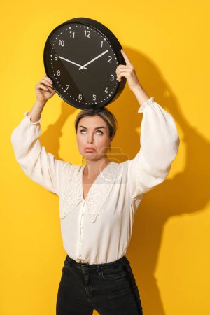 Photo for Upset young woman holding big clock on yellow background - Royalty Free Image