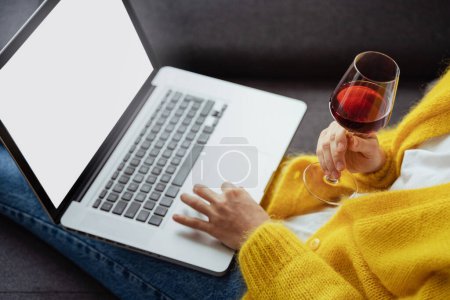 Junge Frau mit einem Glas Rotwein benutzt Laptop zu Hause