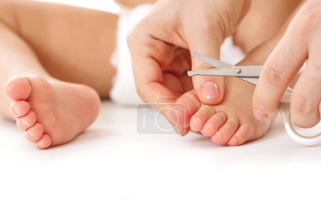 Foto de Primer plano de una madre sosteniendo suavemente el pie de su hijo pequeño y cortando las uñas de los pies sobre un fondo blanco. - Imagen libre de derechos