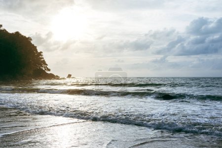 Téléchargez les photos : Vagues de mer mousseuses s'écrasant sur une plage de sable pendant la marée haute avec ciel nuageux en arrière-plan. - en image libre de droit
