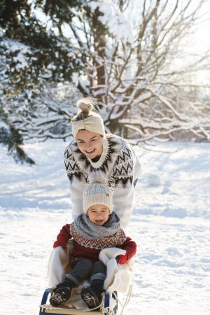 Photo for Mother and her cute little son wearing warm sweaters are having on a sledding hill during sunny winter day - Royalty Free Image