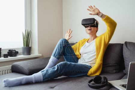 Foto de Mujer joven emocionada sentada en el sofá y usando auriculares de realidad virtual en casa - Imagen libre de derechos