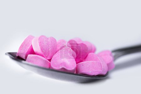 Foto de Primer plano de una cuchara metálica llena de píldoras en forma de corazón rosa sobre fondo blanco. - Imagen libre de derechos