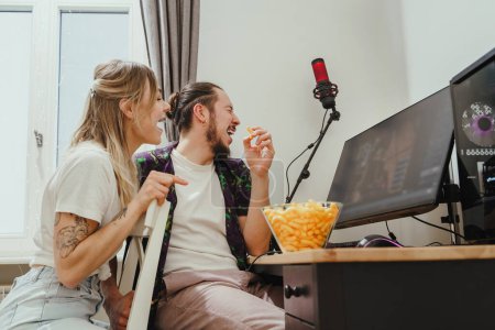 Photo for Young couple eating cheese puffs while playing video games or watching something online at home - Royalty Free Image