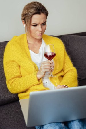 Foto de Mujer joven sentada en el sofá bebiendo vino tinto mientras trabaja en su computadora portátil en casa - Imagen libre de derechos