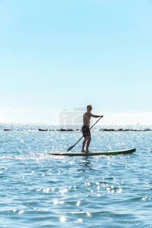 Photo for Young male surfer is riding a standup paddleboard and rowing with a paddle in ocean. - Royalty Free Image